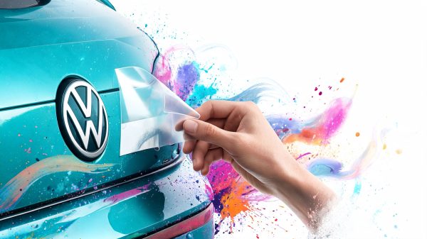 Hand peeling a clear film off the front of a teal Volkswagen car, with colorful splash paint background