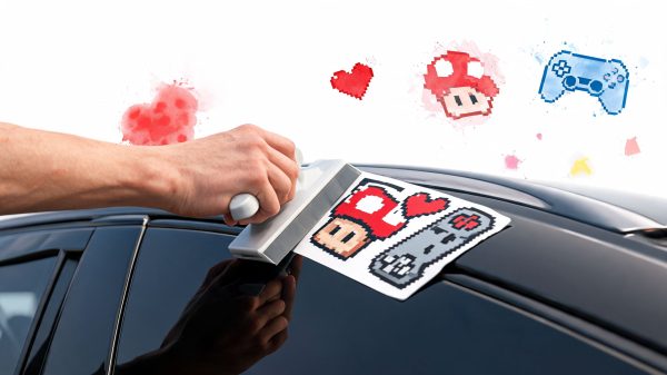 Person applies a retro pixel-art sticker to a car window, featuring a Mario-style character, red hearts, and a blue game controller.